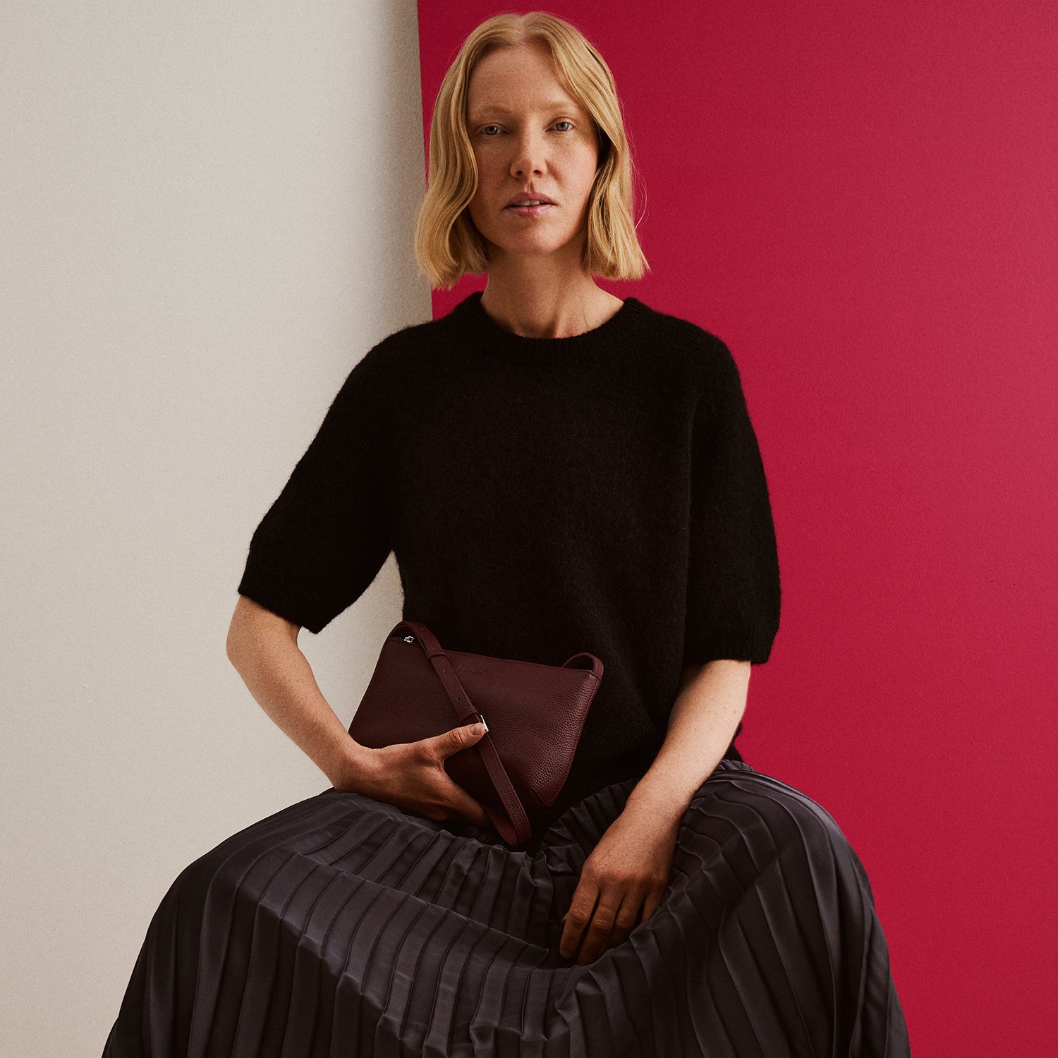 Nanson naisten musta lyhythihainen Tuohi neulepusero ja musta Taite hame mallin päällä, kädessä viininpunainen Naru olkalaukku.