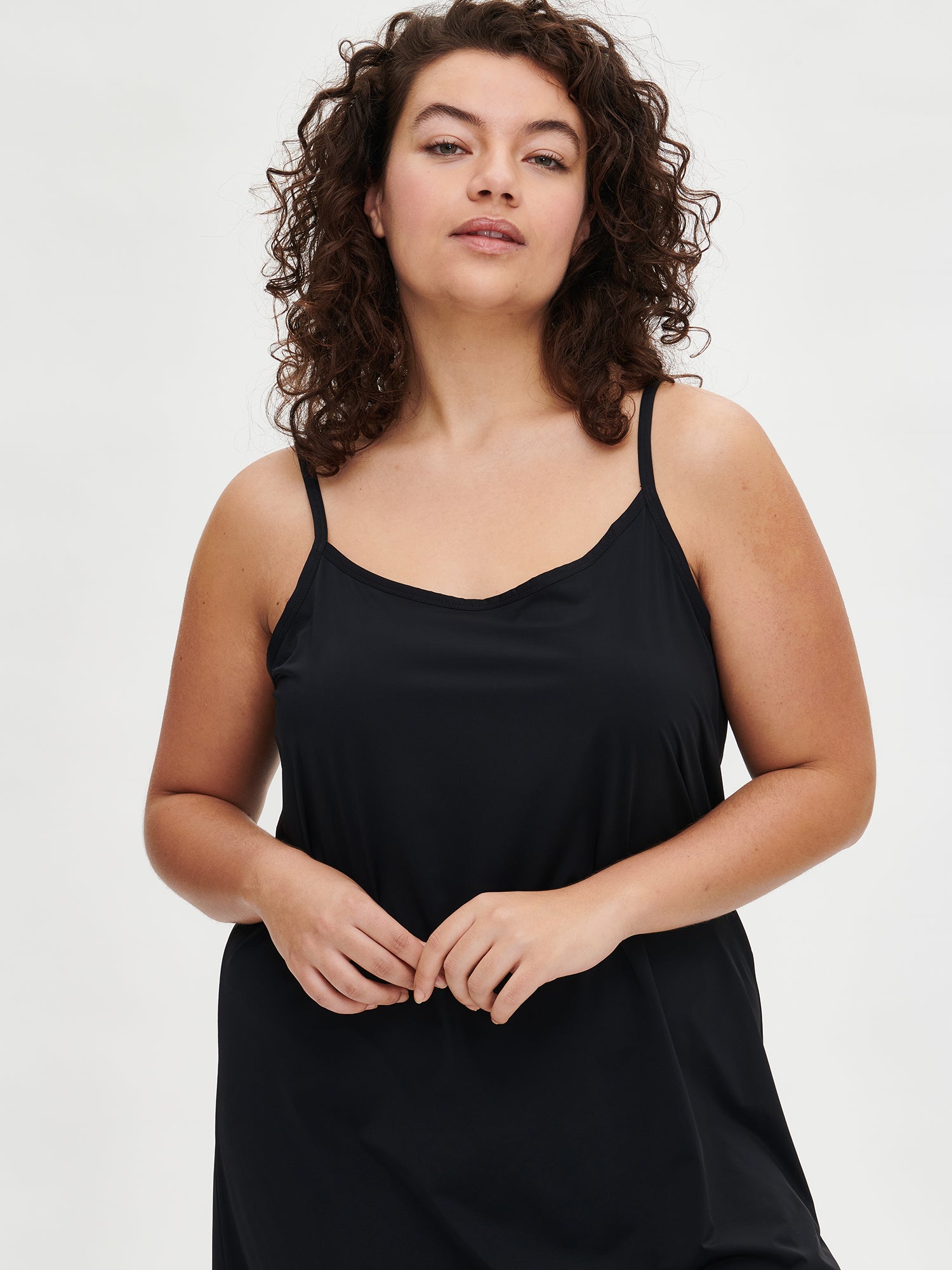 Nanson naisten musta Basic-alusmekko ohuilla olkaimilla, puolipitkä edestä kuvattu näkymä plus size-mallin päällä. Mallin kädet yhdessä.