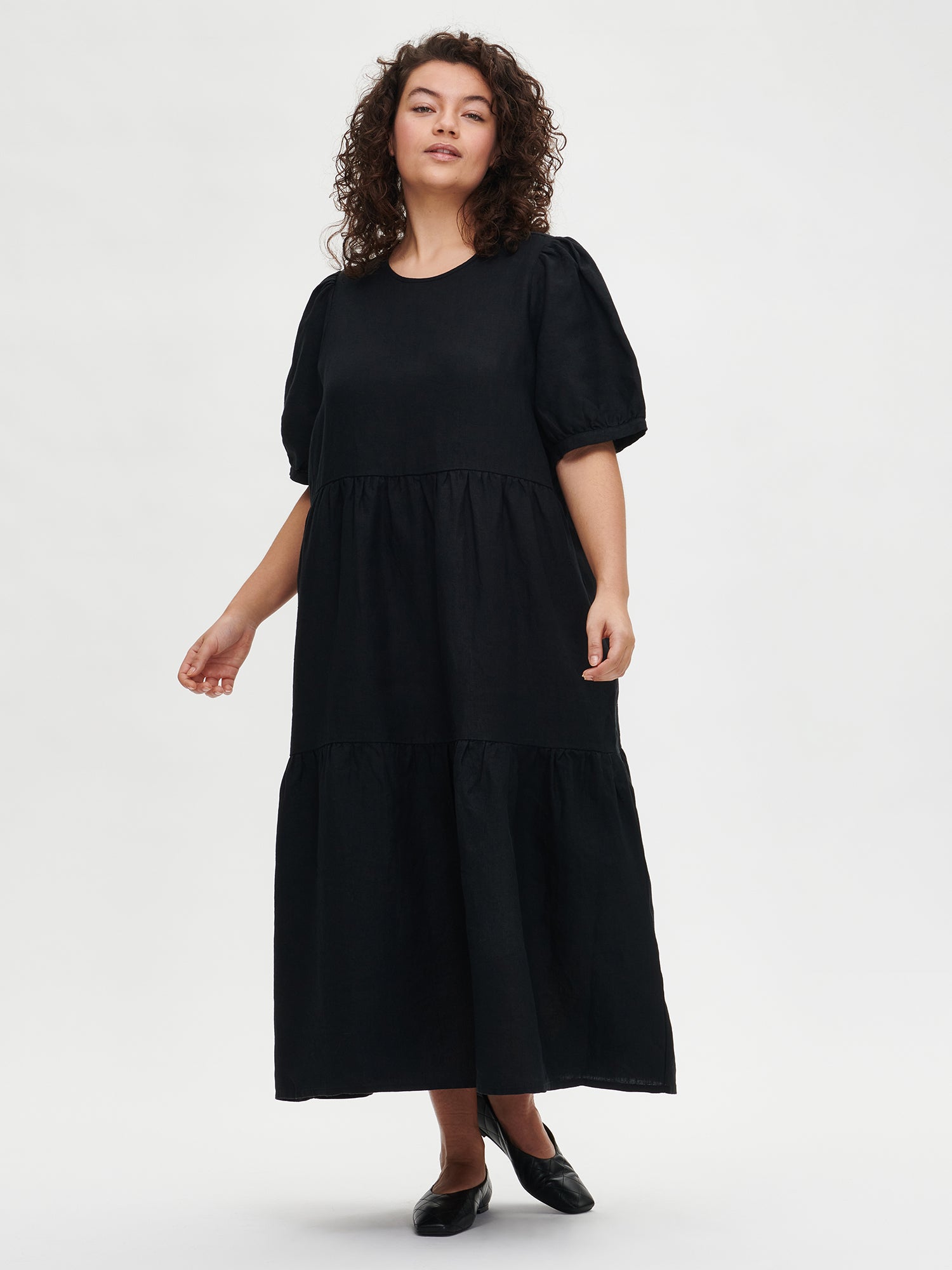 Nanson naisten kaunis musta Sarastus-pellavamekko volyymisilla frillakerroksilla ja lyhyillä puhvihihoilla mallin päällä koossa XL.