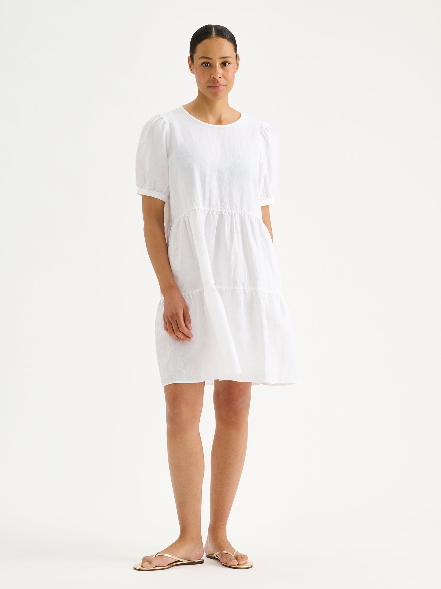 Naisten ajaton, lyhyt valkoinen Sarastus-pellavamekko volyymisilla frillakerroksilla ja lyhyillä puhvihihoilla, joissa on napit säätövaralla mallin päällä sandaalien kanssa.