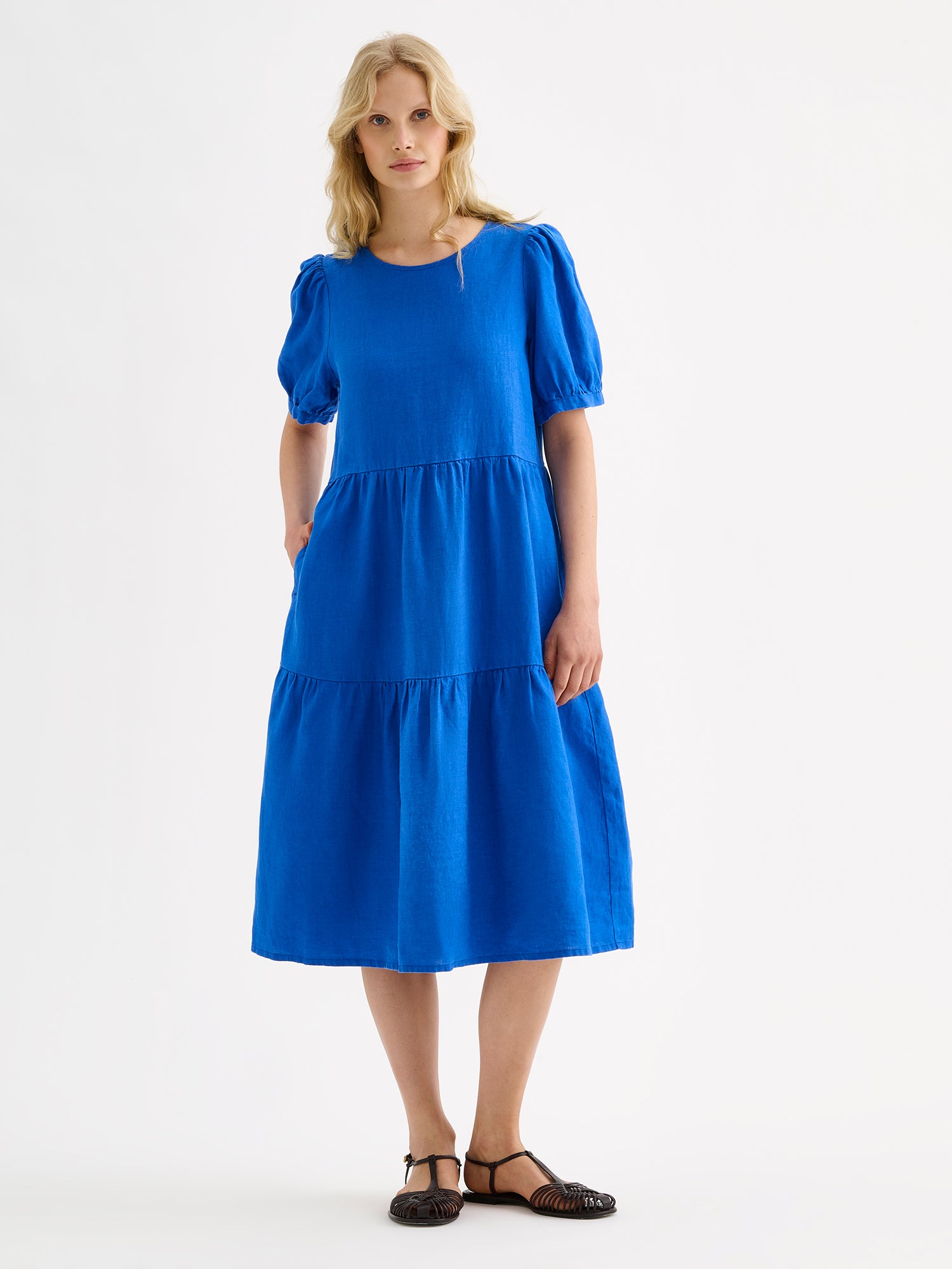 Nanson naisten kaunis sininen midimittainen Sarastus-pellavamekko volyymisilla frillakerroksilla ja lyhyillä puhvihihoilla, vaaleahiuksisen mallin päällä.