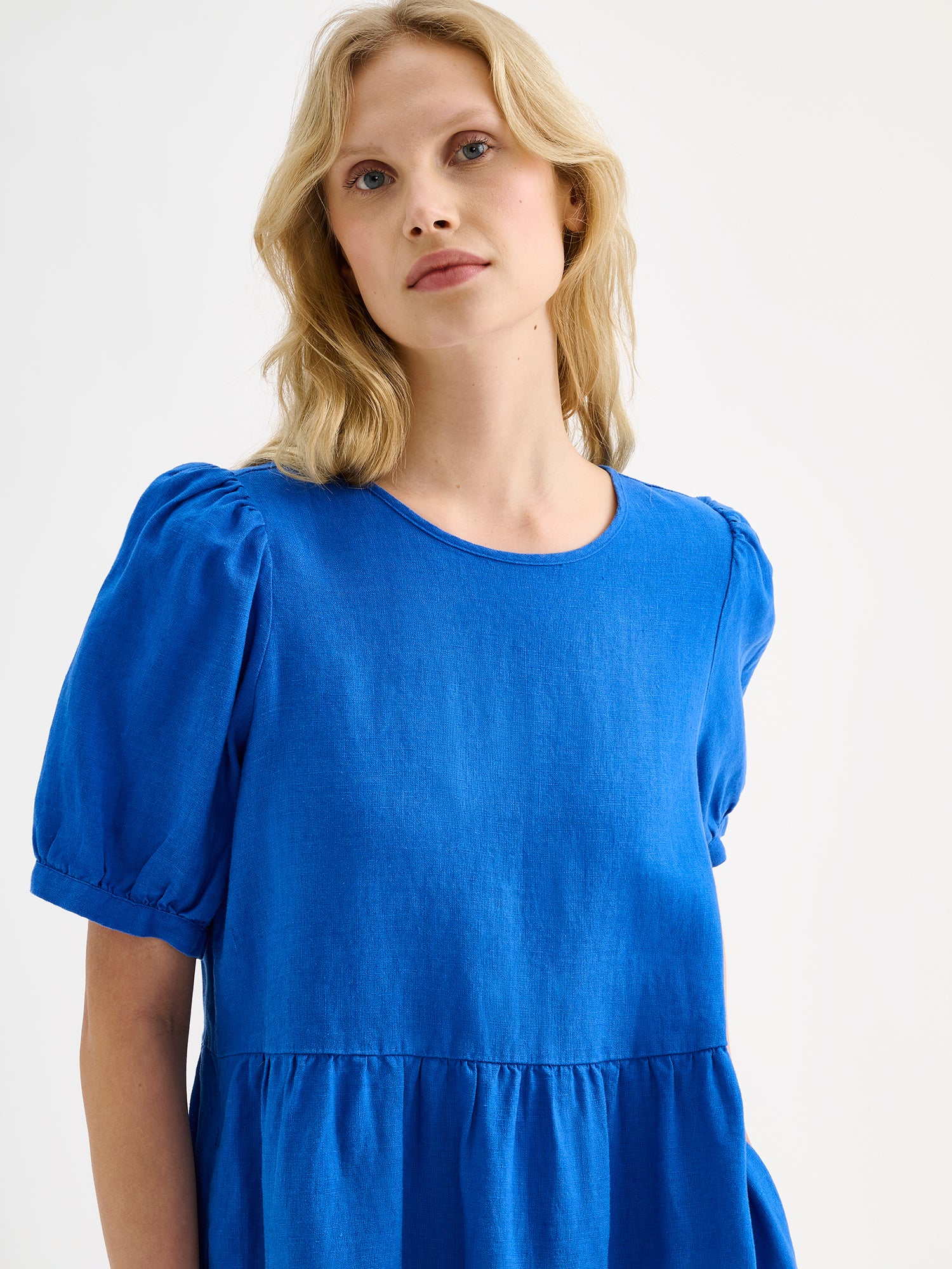Nanson naisten kaunis sininen midimittainen Sarastus-pellavamekko volyymisilla frillakerroksilla ja lyhyillä puhvihihoilla, vaaleahiuksisen mallin päällä lähikuvassa.