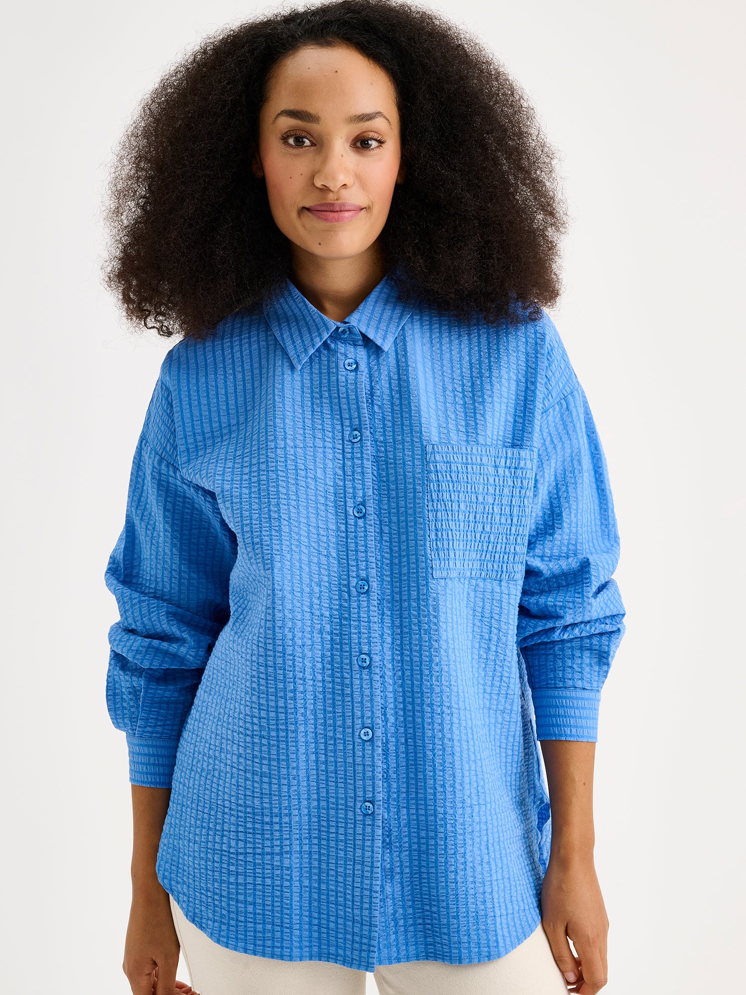 Nanson naisten tyylikäs sininen Maari-paitapusero, jossa kaulus, pudotettu olkalinja, rintatasku ja napit edessä. Kevyessä kudotussa materiaalissa on kaunis pintastruktuuri. Lähikuvassa mallin päällä vaaleiden farkkujen kanssa.