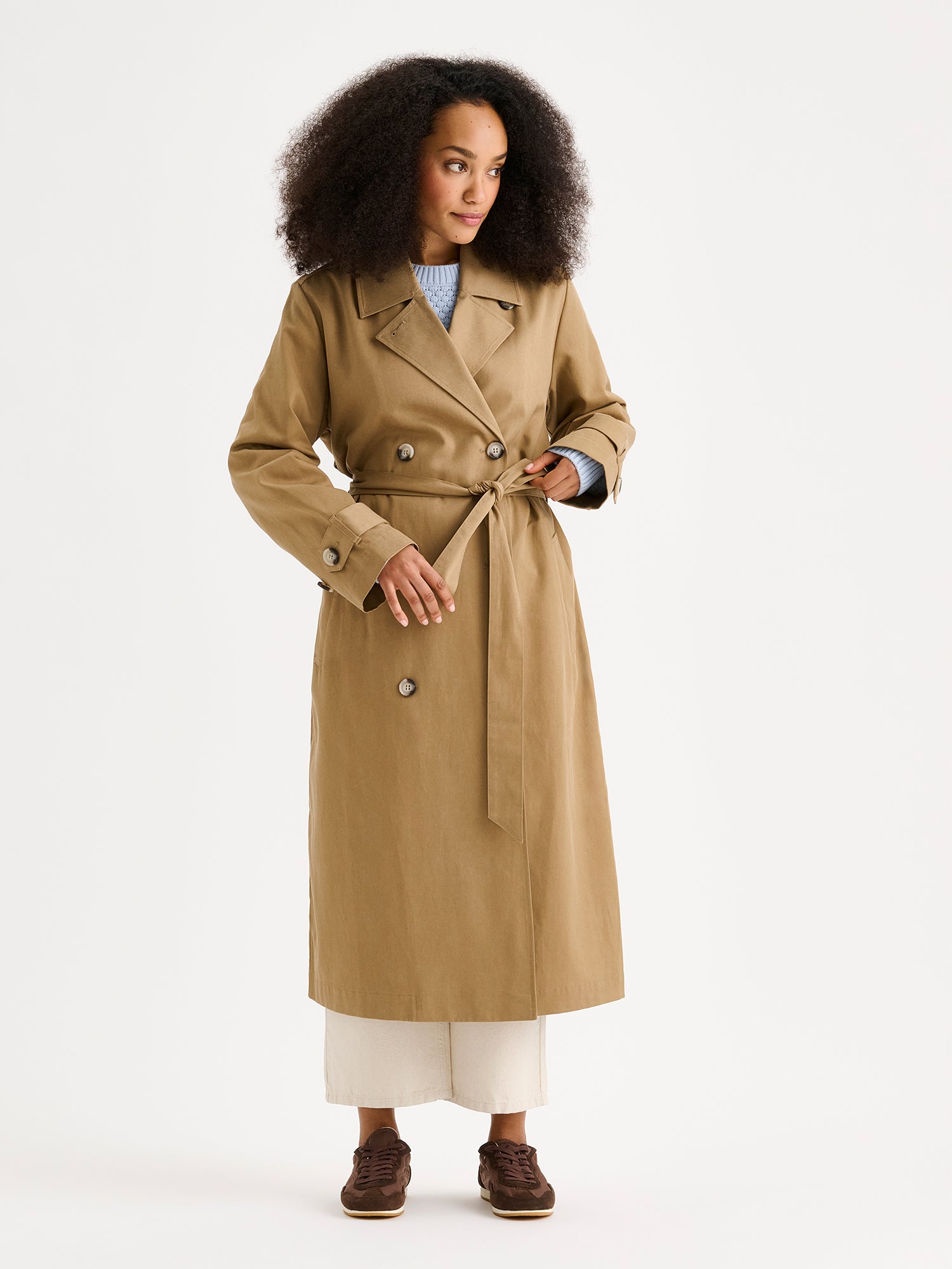 Naisten klassisen tyylikäs ruskea Klaara-trenssitakki mallin päällä vaaleiden farkkujen ja tennareiden kanssa.