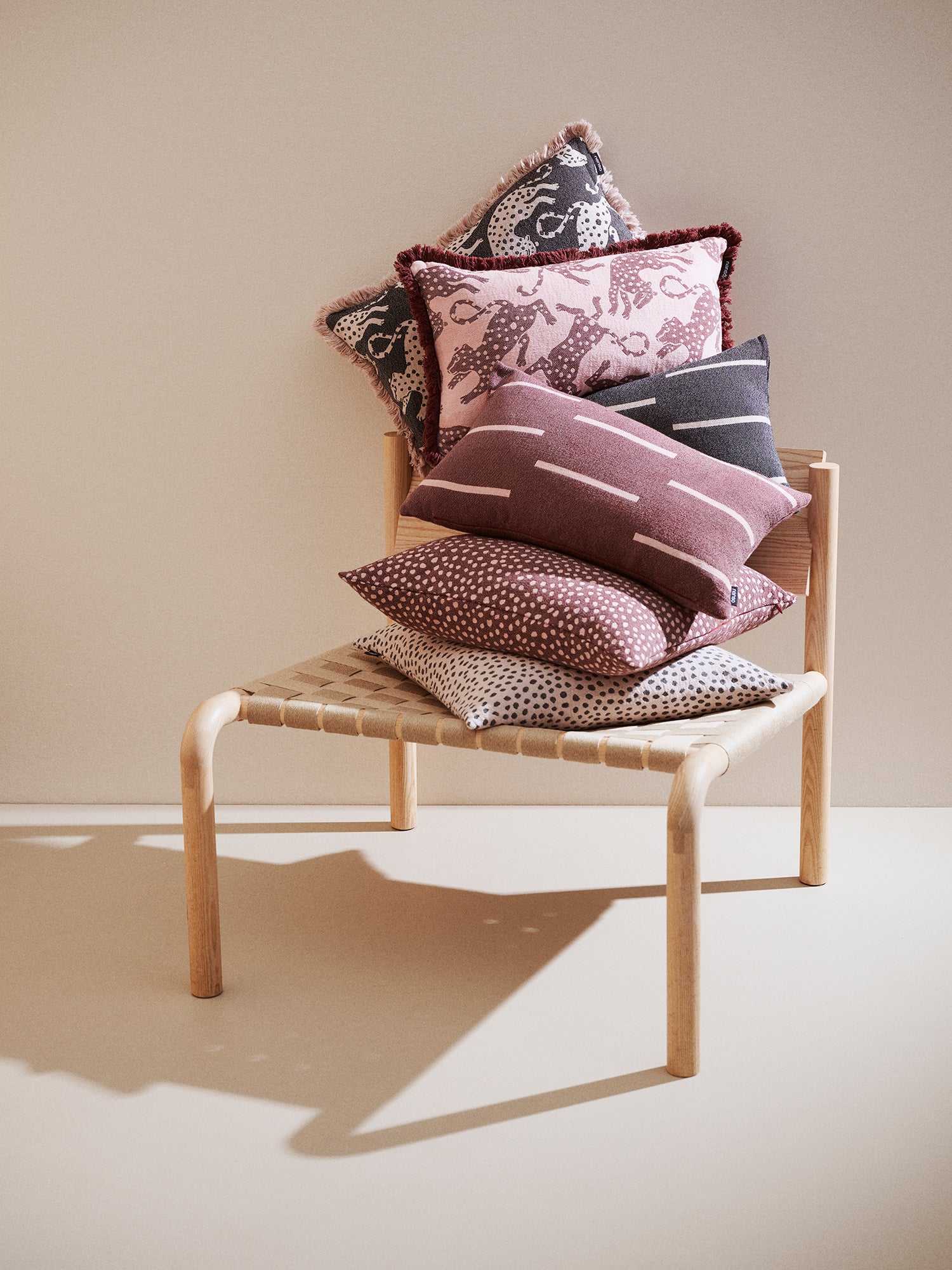 Vaaleanpunainen Hapsu-tyynynpäällinen kuvattuna mustan sävyisen Hapsun kanssa sekä muiden saman sävyisten koristetyynyjen kanssa tuolin päällä.