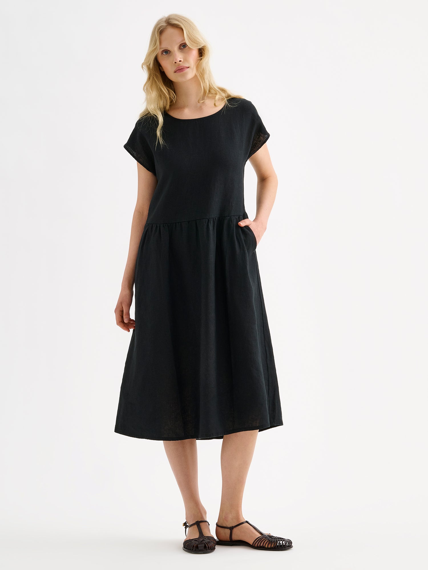 Nanson naisten rento midipituinen lyhythihainen musta Saari-pellavamekko taskuilla, rypytetyllä helmalla ja ovaalin mallisella pääntiellä mallin päällä sandaalien kanssa.