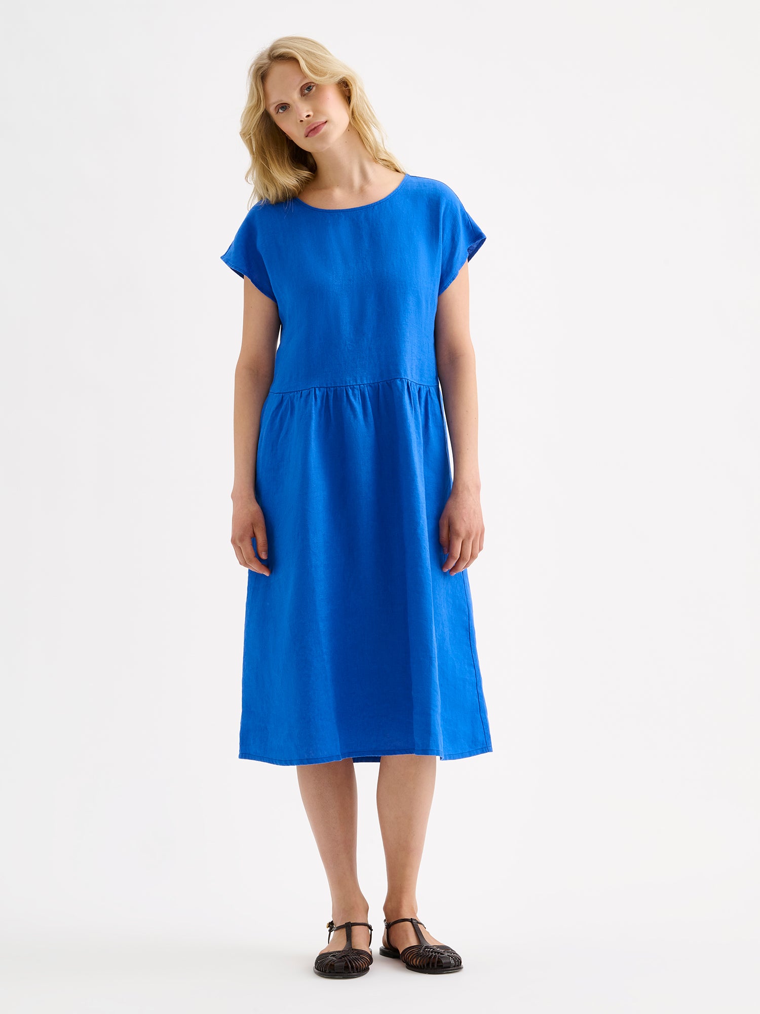 Nanson naisten rento midipituinen lyhythihainen sininen Saari-pellavamekko taskuilla, rypytetyllä helmalla ja ovaalin mallisella pääntiellä mallin päällä sandaalien kanssa.
