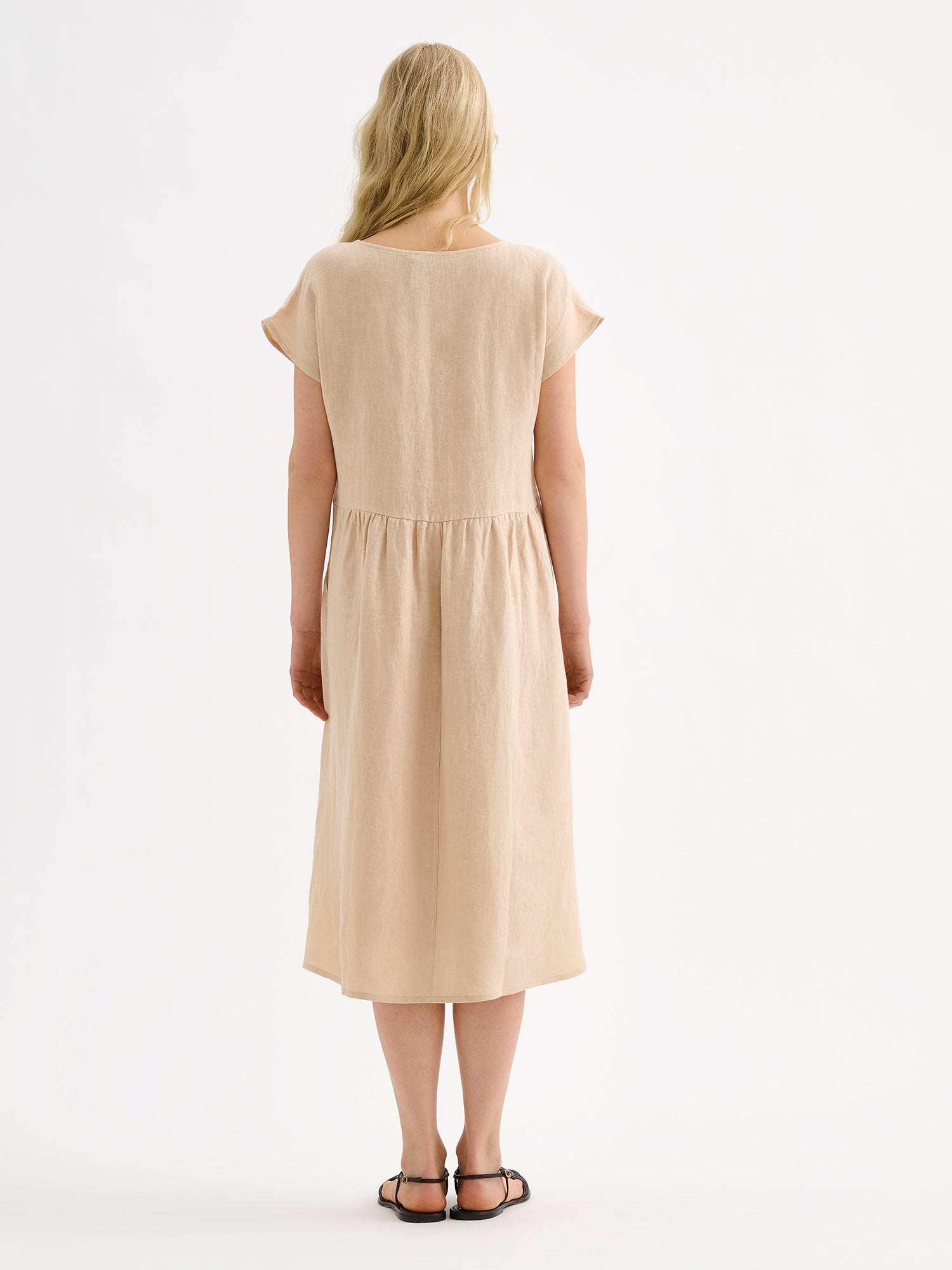 Nanson naisten rento midipituinen lyhythihainen beige Saari-pellavamekko taskuilla, rypytetyllä helmalla ja ovaalin mallisella pääntiellä mallin päällä sandaalien kanssa takaa kuvattuna.
