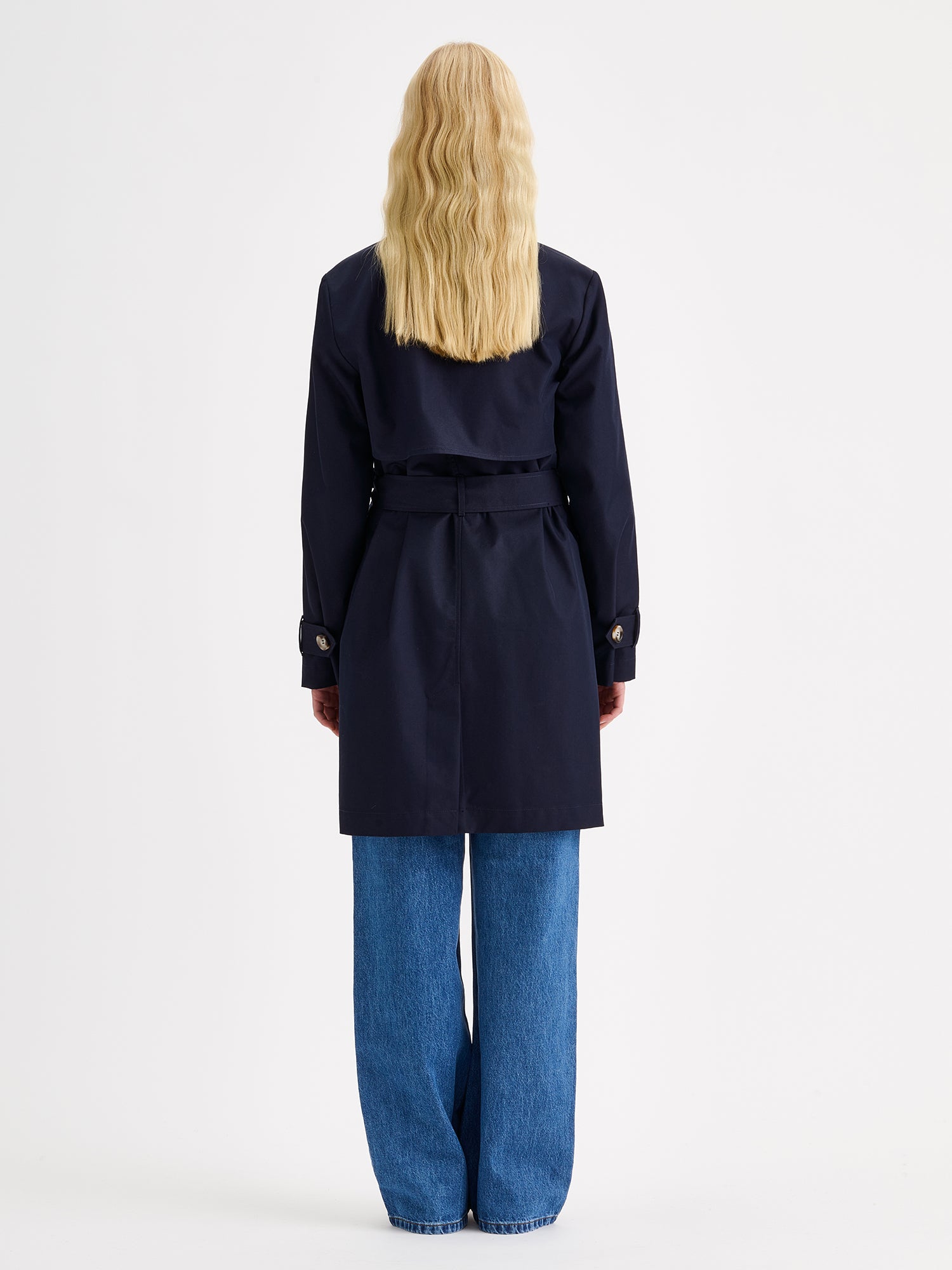 Nanson naisten tummansininen Midi Trenssi-takki vaaleahiuksisen mallin päällä farkkujen ja tennareiden kanssa, kuva otettu takaa päin.