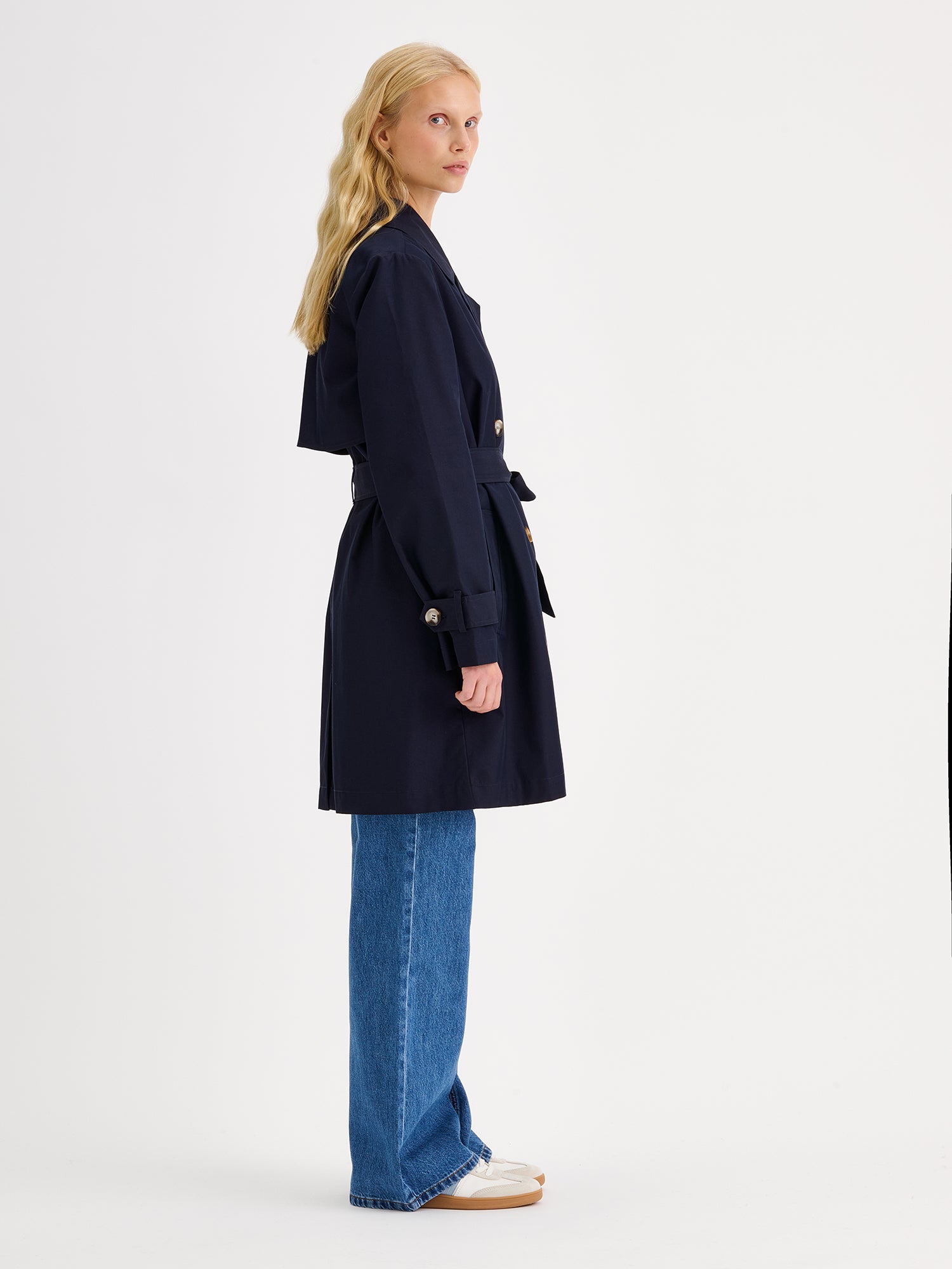 Nanson naisten tummansininen Midi Trenssi-takki vaaleahiuksisen mallin päällä farkkujen ja tennareiden kanssa sivusta kuvattuna.