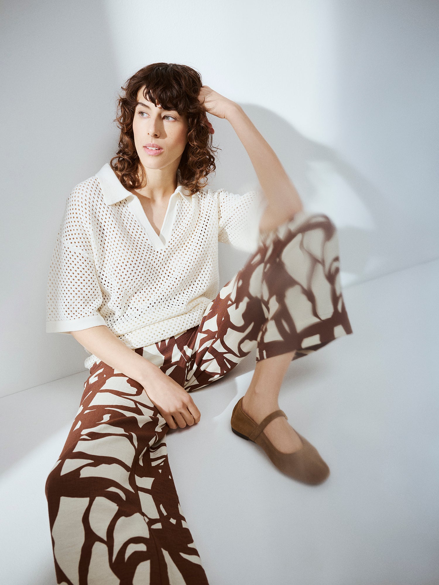 Nanson naisten luonnonvalkoinen Aure -reikäneulepusero lattialla istuvan mallin päällä kuosillisten Palsta-housujen kanssa.