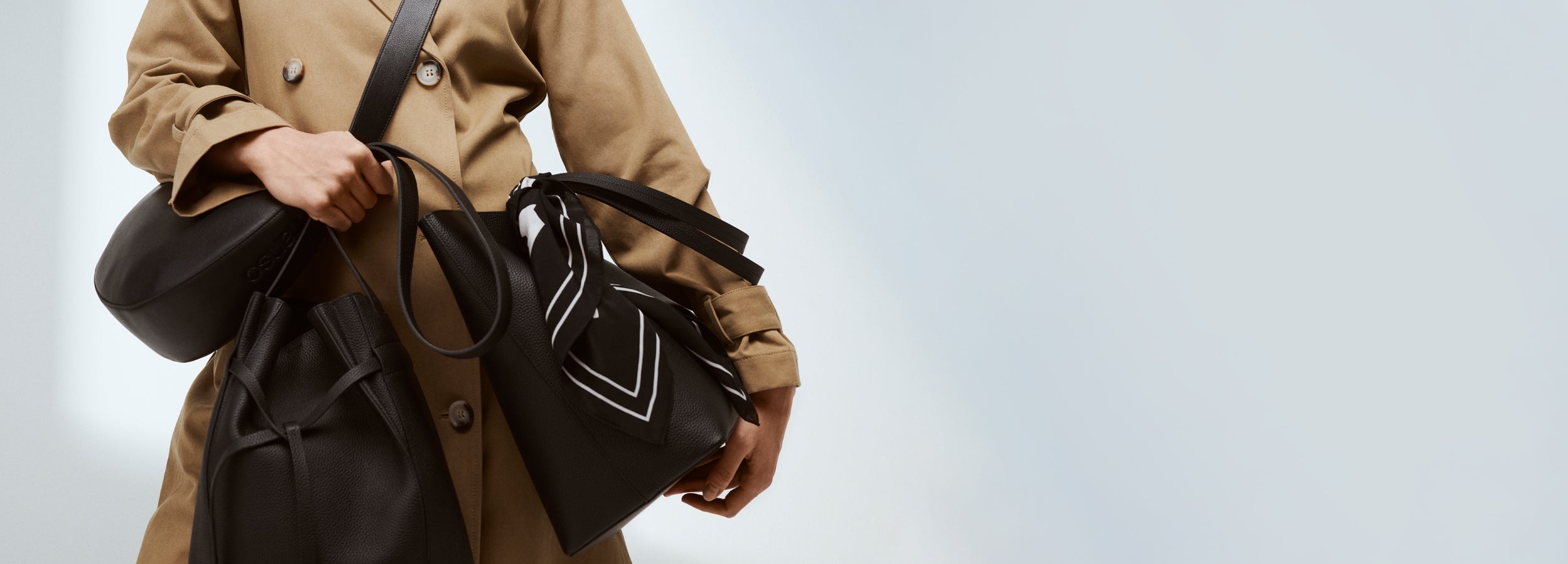 Nanson mustat nahkaiset laukut mallin olalla ja kädessä Klaara trenssitakin kanssa läheltä kuvattuna. Maxi Naru -laukkuun sidottu mustavalkoinen Palsta -huivi koristeeksi