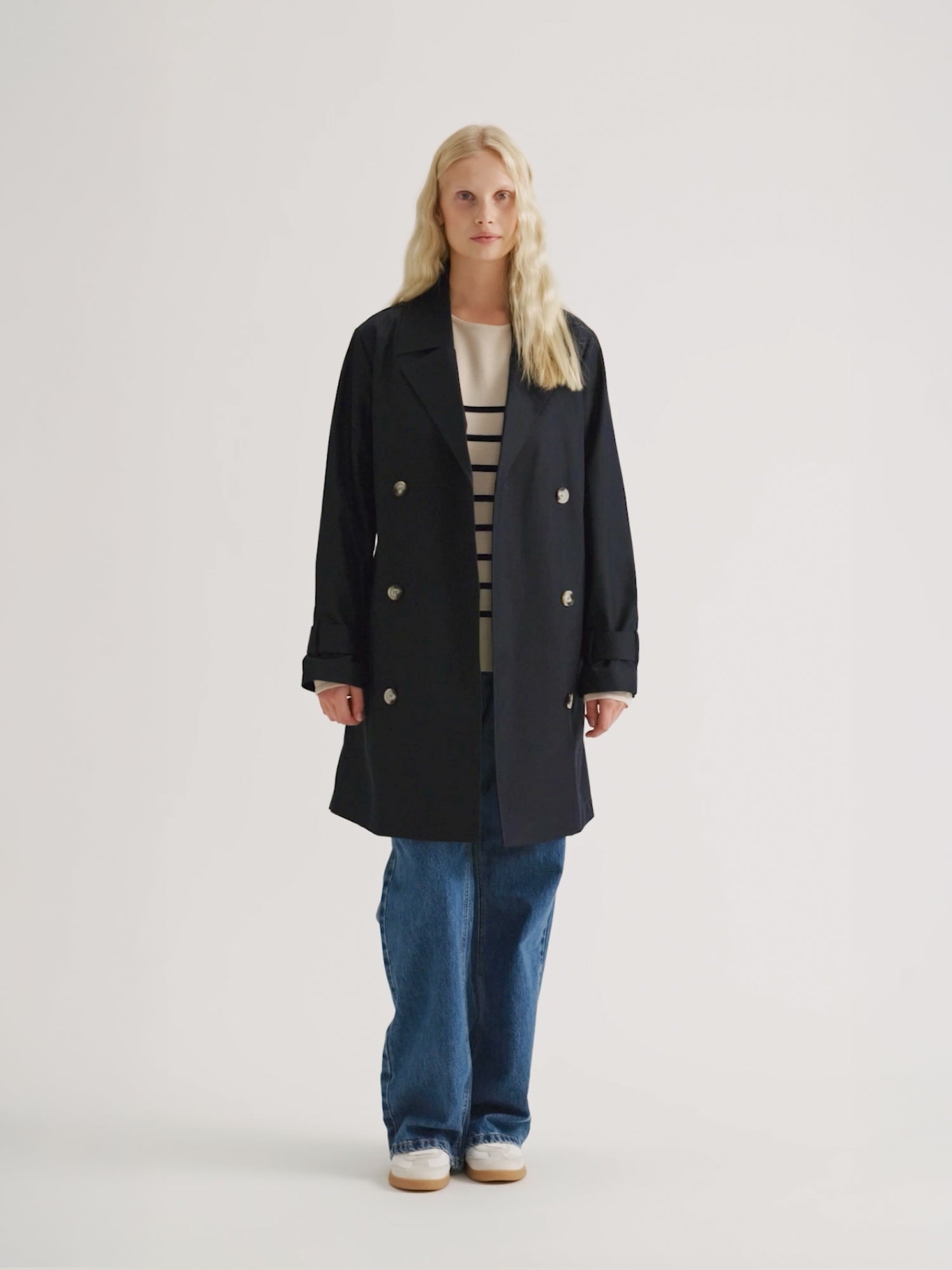 Nanson naisten tummansininen Midi Trenssi-takki vaaleahiuksisen mallin päällä farkkujen ja tennareiden kanssa, takki auki.