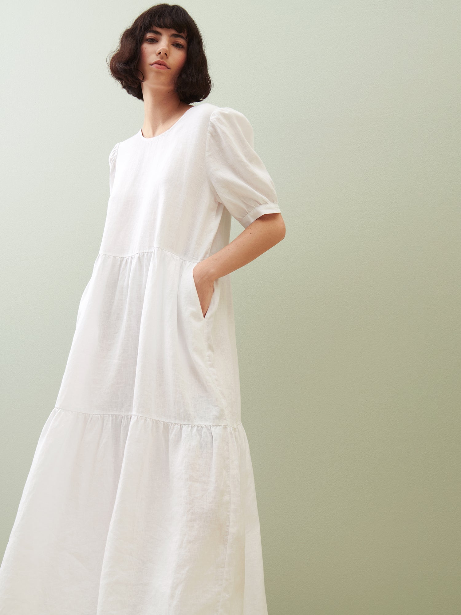 Nanson naisten kaunis valkoinen Sarastus-mekko volyymisilla frillakerroksilla ja lyhyillä puhvihihoilla vihreällä taustalla.