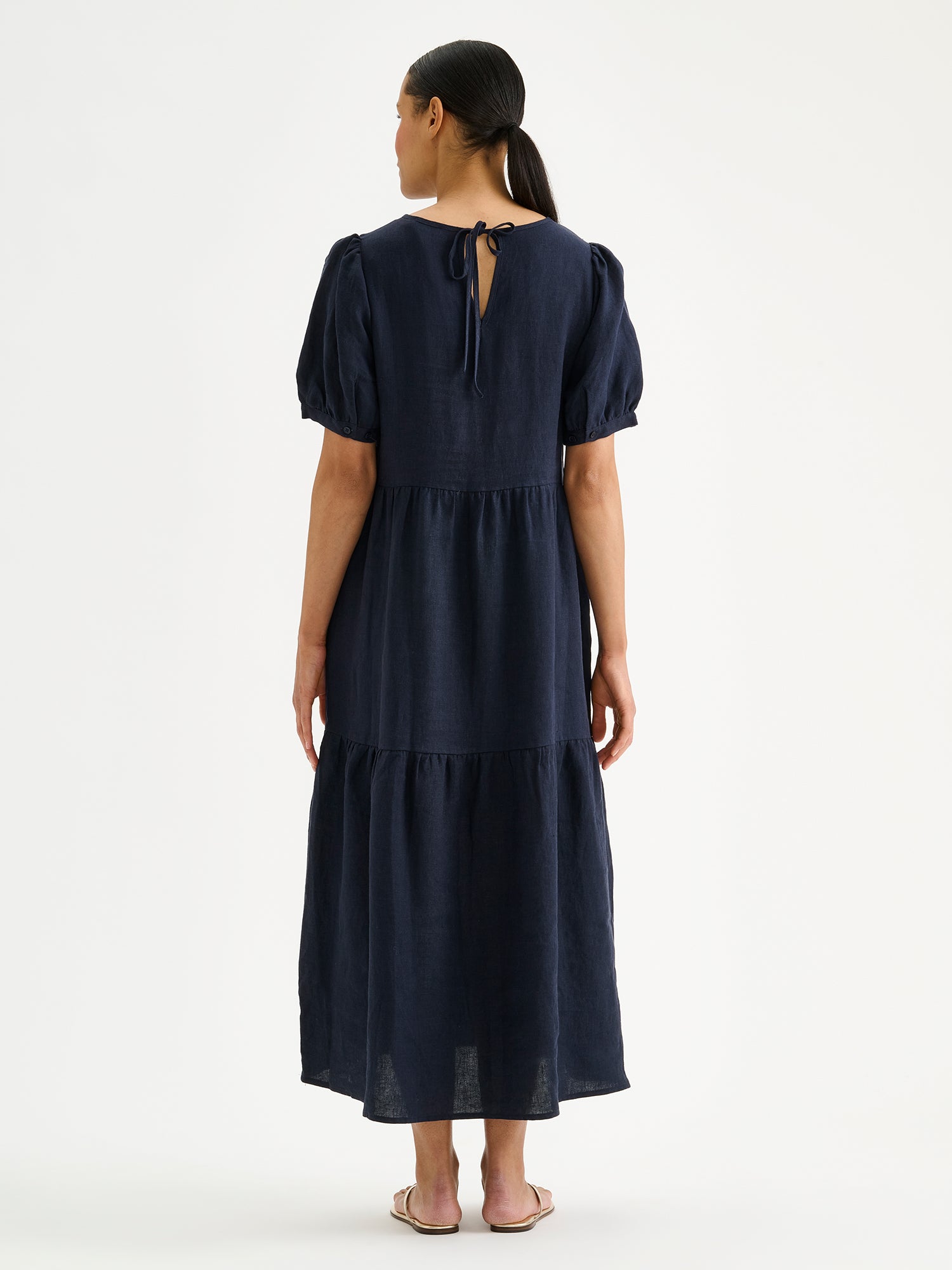 Naisten kaunis tummansininen Sarastus-pellavamekko volyymisilla frillakerroksilla ja lyhyillä puhvihihoilla mallin päällä sandaalien kanssa takaa kuvattuna.