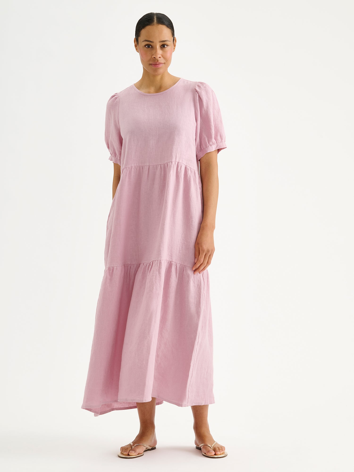 Naisten kaunis roosa Sarastus-pellavamekko volyymisilla frillakerroksilla ja lyhyillä puhvihihoilla mallin päällä sandaalien kanssa.