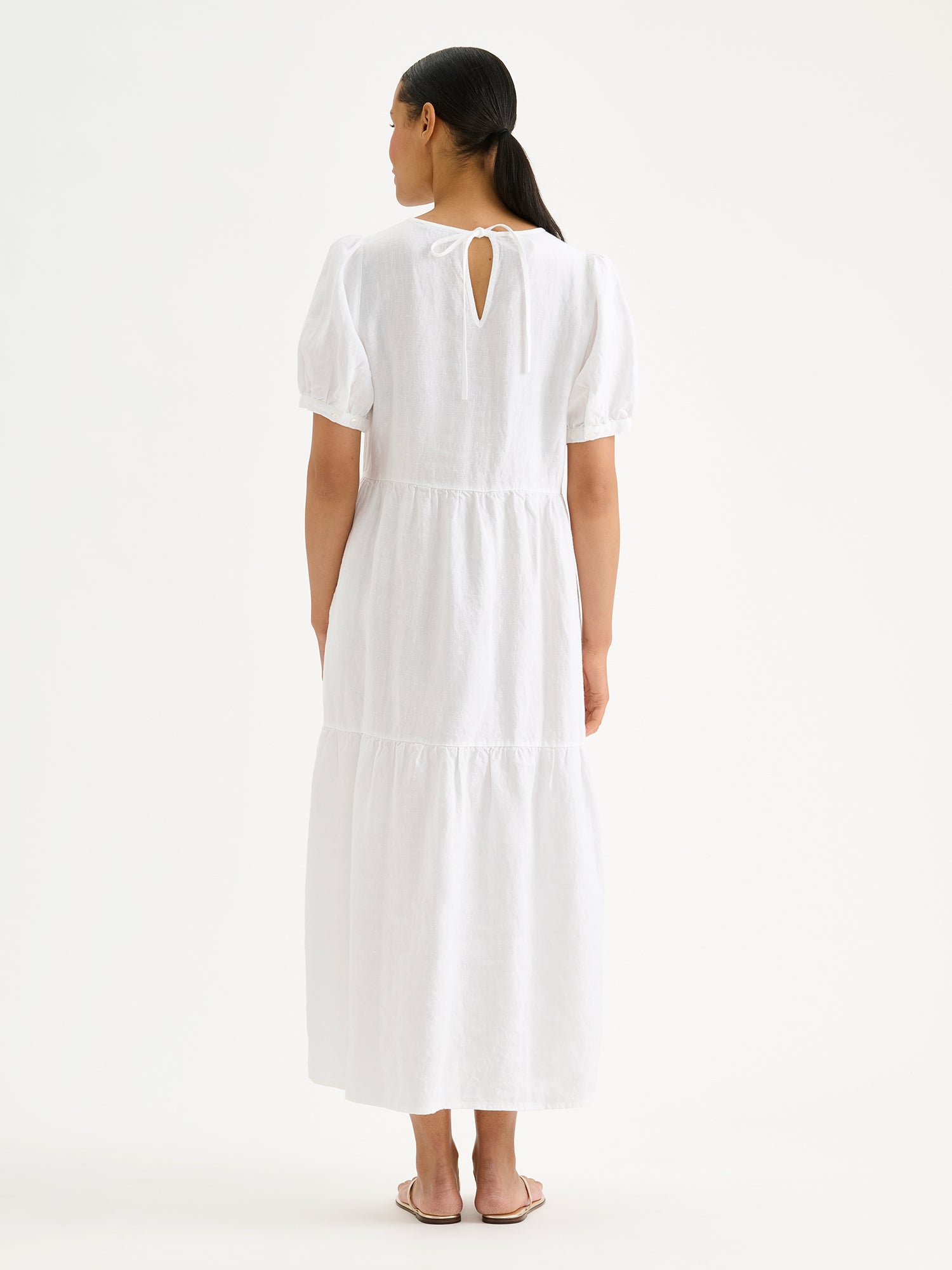 Naisten kaunis valkoinen Sarastus-pellavamekko volyymisilla frillakerroksilla ja lyhyillä puhvihihoilla mallin päällä takaa kuvattuna