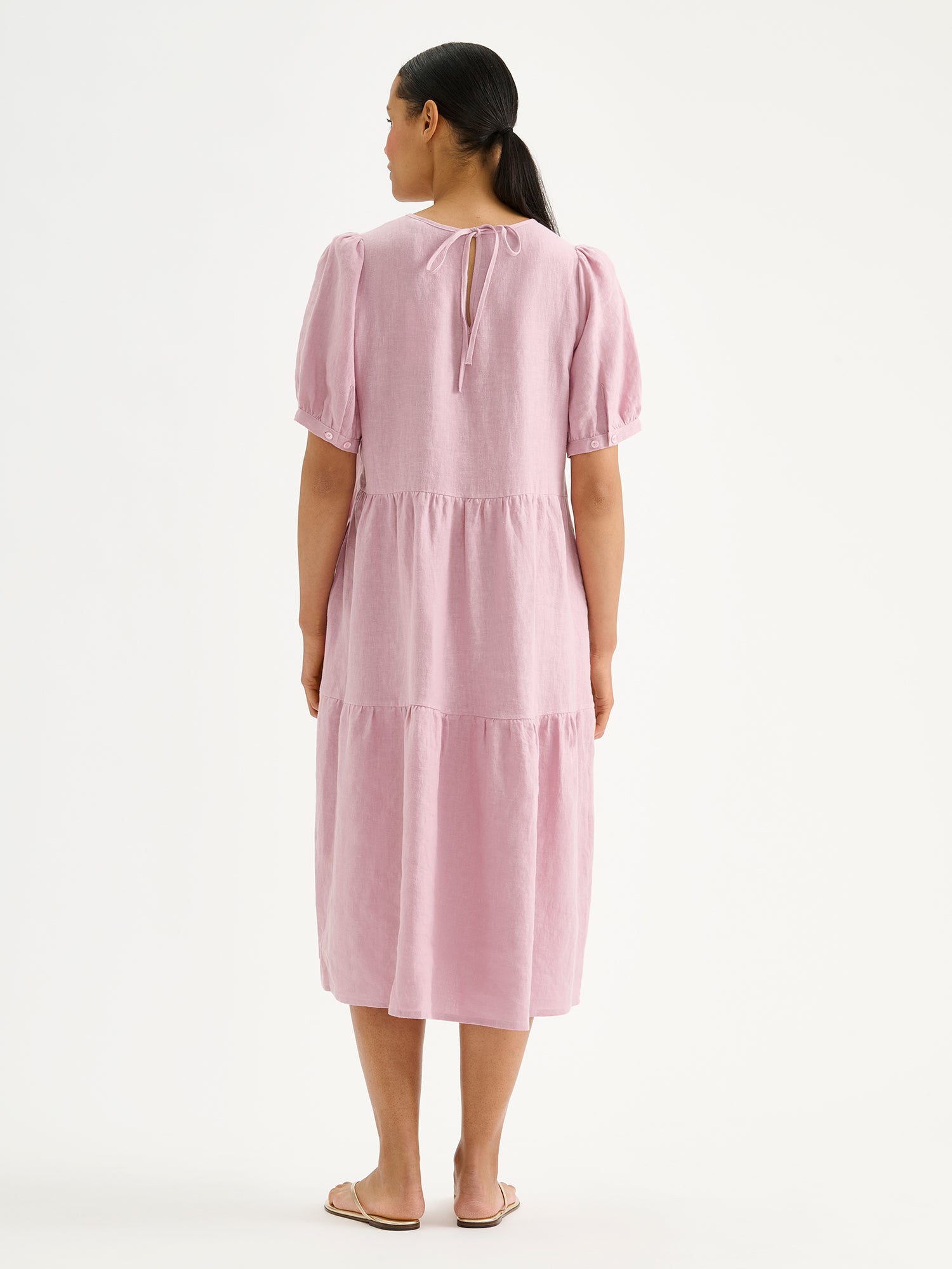 Nanson naisten kaunis roosa midimittainen Sarastus-pellavamekko volyymisilla frillakerroksilla ja lyhyillä puhvihihoilla mallin päällä sandaalien kanssa takaa päin kuvattuna.