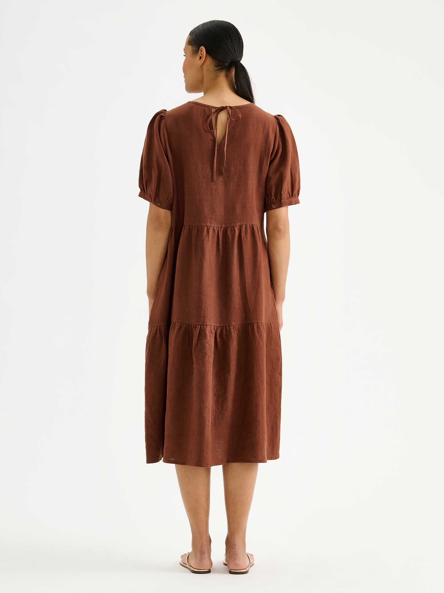 Nanson naisten kaunis ruskea midimittainen Sarastus-pellavamekko volyymisilla frillakerroksilla ja lyhyillä puhvihihoilla mallin päällä sandaalien kanssa takaa kuvattuna.