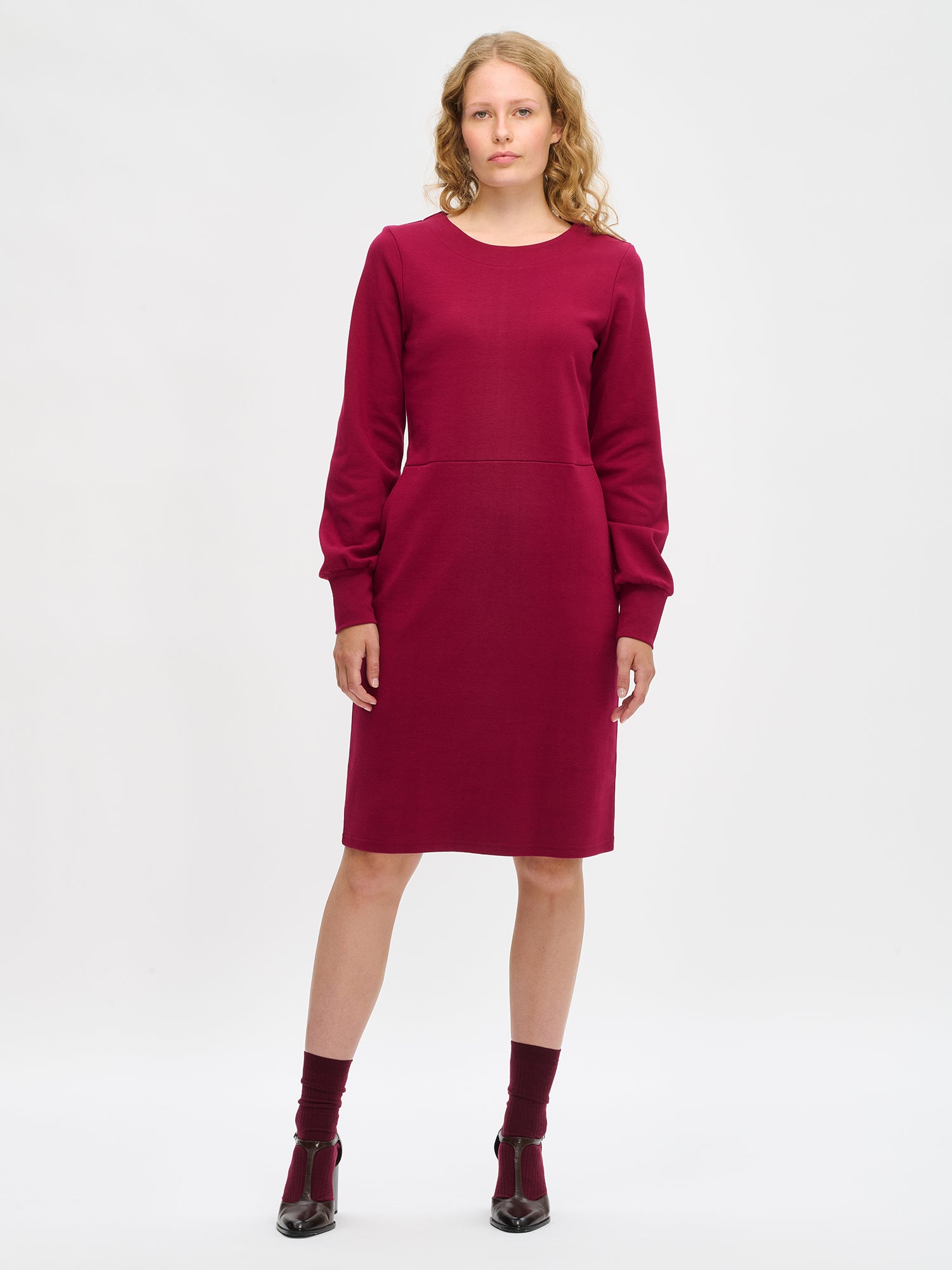 Nanson naisten viininpunainen Pauliina-mekko mallin yllä kokokuvassa, polvipituinen, pitkät hihat poimuttuvat leveän hihansuun yläpuolelle, venepääntie ja istuva vyötärö.