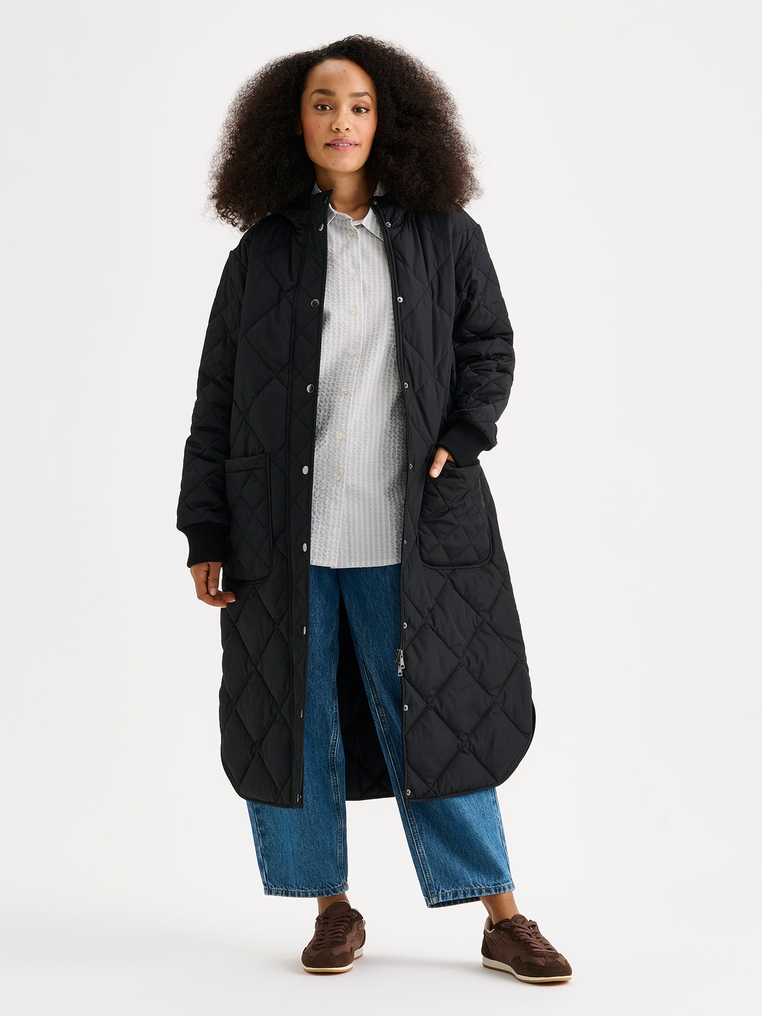 Nanson naisten rennon tyylikäs musta Maxi Hood-tikkitakki kevyellä vanulla ja lämpimällä hupulla. Hupullinen tikkitakki on täydellinen valinta välikauteen. Mallin päällä, takki auki.