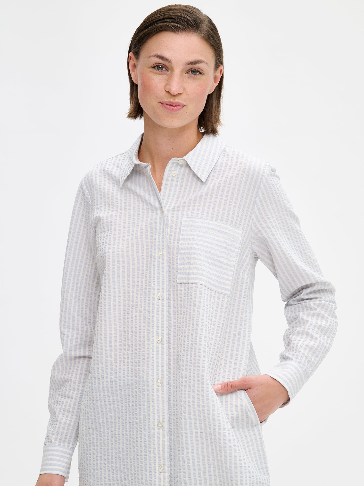 Nanson naisten raidallinen vaalean sininen Marea-paitamekko kauniilla pintastruktuurilla ja klassisella kauluksella. Mekossa on napitus, sivutaskut ja pyöristetty helma. Mallin päällä lähikuvassa.