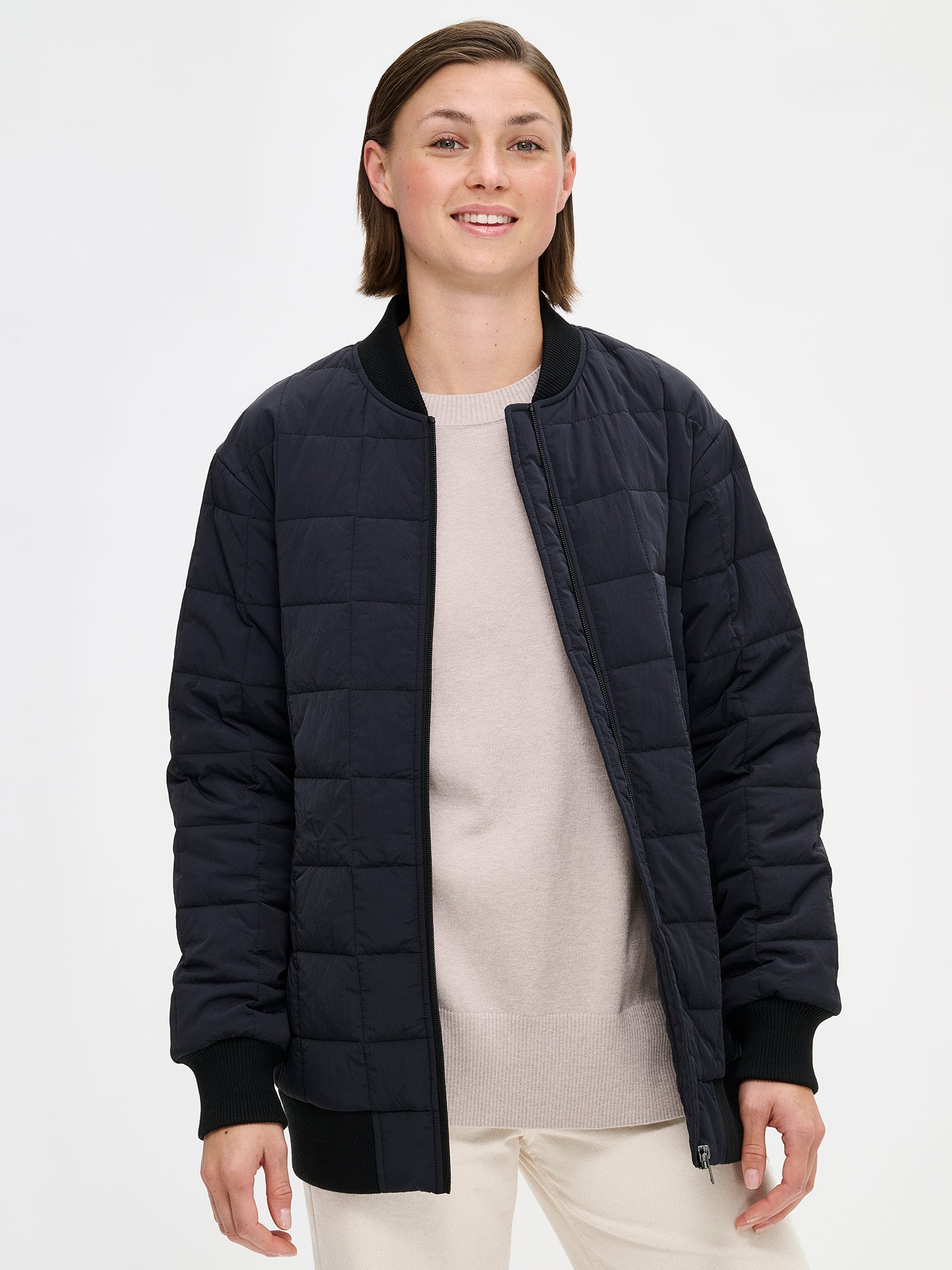 Nanson naisten rento Merje bomber tikkitakki kevyellä vanulla. Takin pääntiellä, hihansuissa ja helmassa on neulosresori. Edessä on vetoketju ja taskut. Takin olkalinja on pudotettu. Mallin päällä auki kuvattuna valkoisten farkkujen kanssa läheltä.