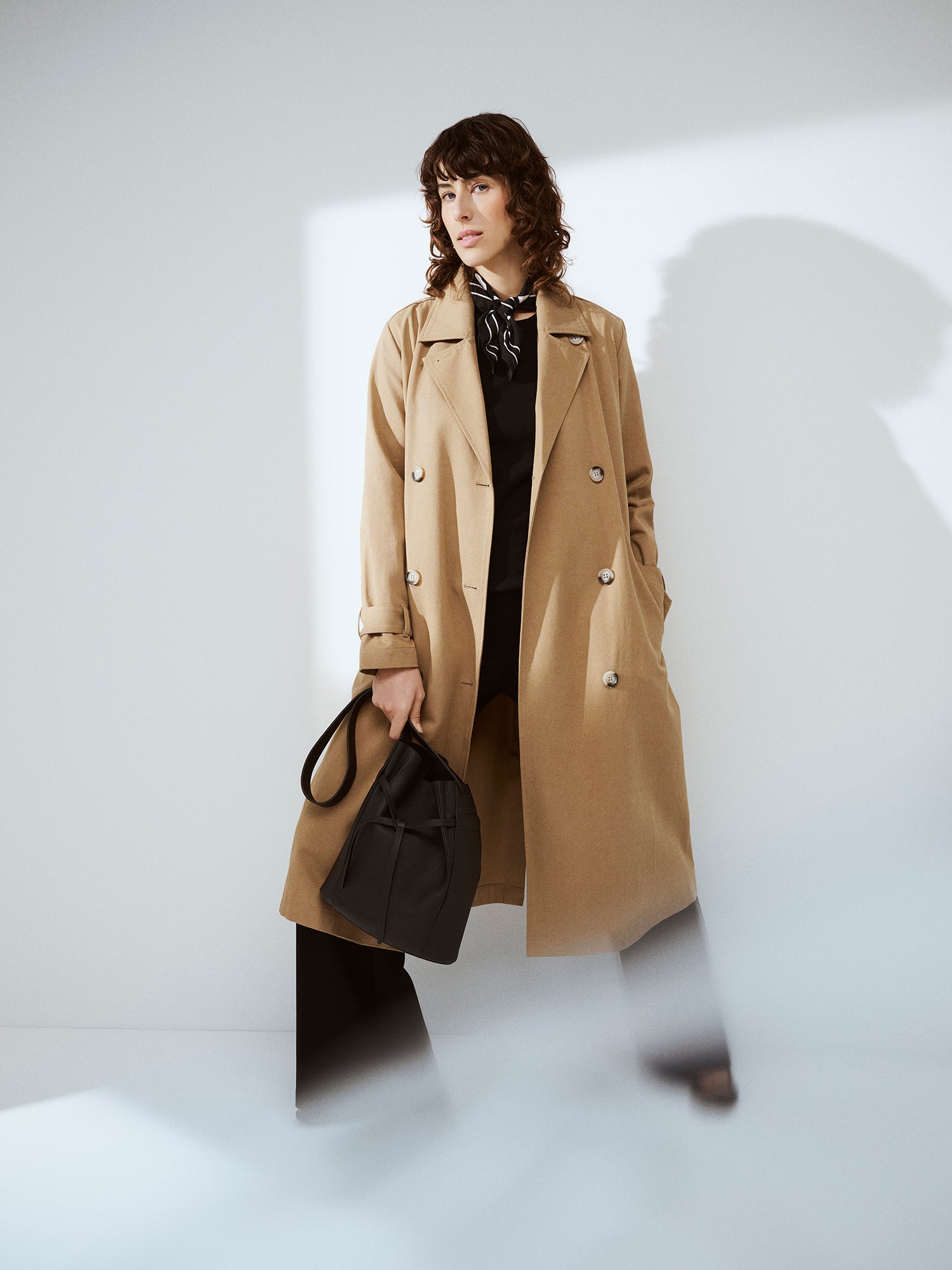 Naisten klassisen tyylikäs ruskea Klaara-trenssitakki mallin päällä mustan asun ja nahkalaukun kanssa.