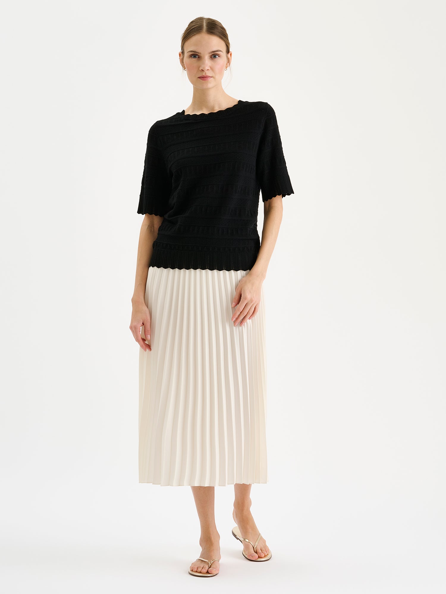 Naisten kauniisti laskeutuva valkoinen pliseerattu Ariel-hame mallin päällä mustan Koru-neulepuseron kanssa.