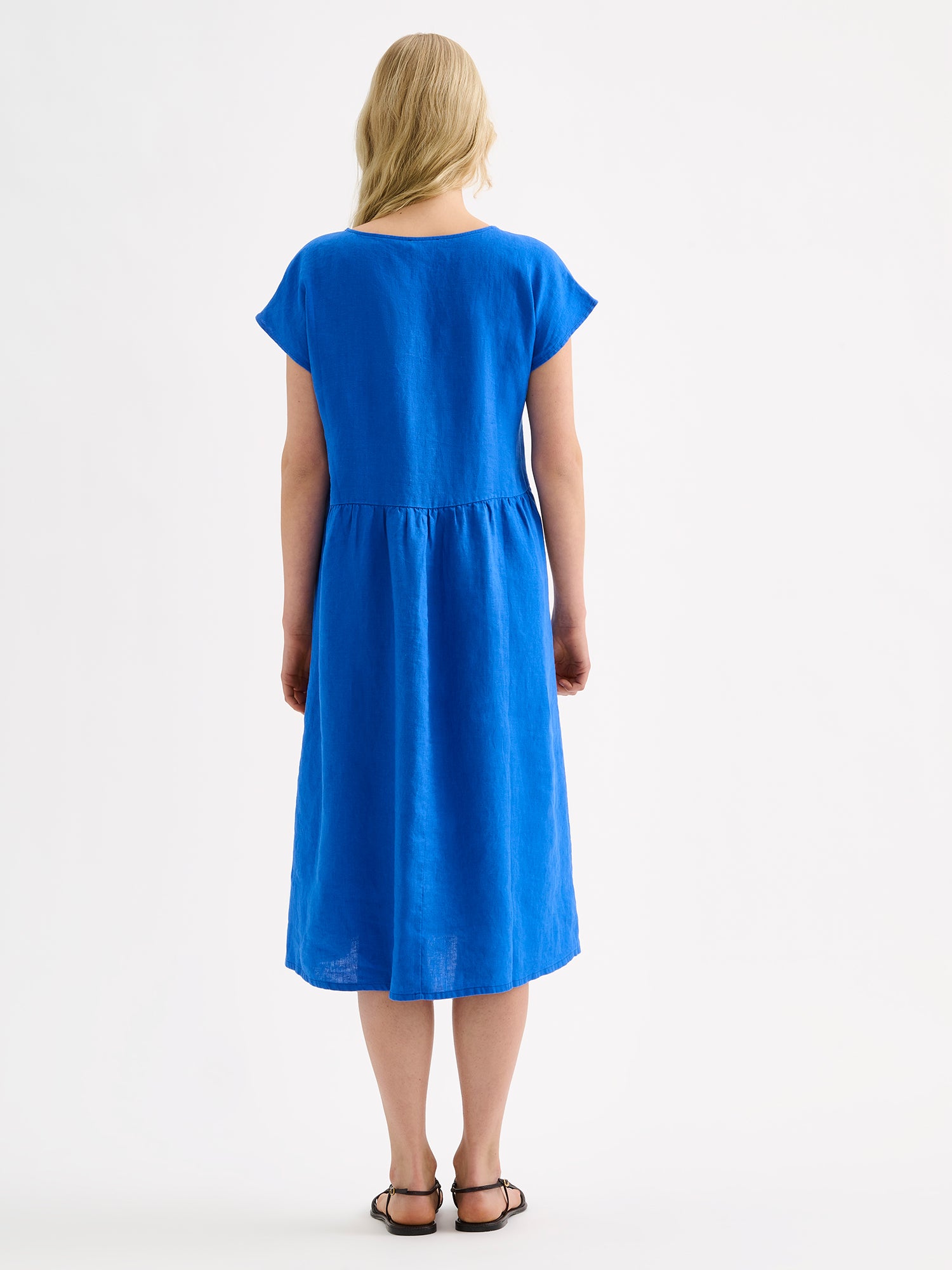 Nanson naisten rento midipituinen lyhythihainen sininen Saari-pellavamekko taskuilla, rypytetyllä helmalla ja ovaalin mallisella pääntiellä mallin päällä sandaalien kanssa, takaa kuvattuna.