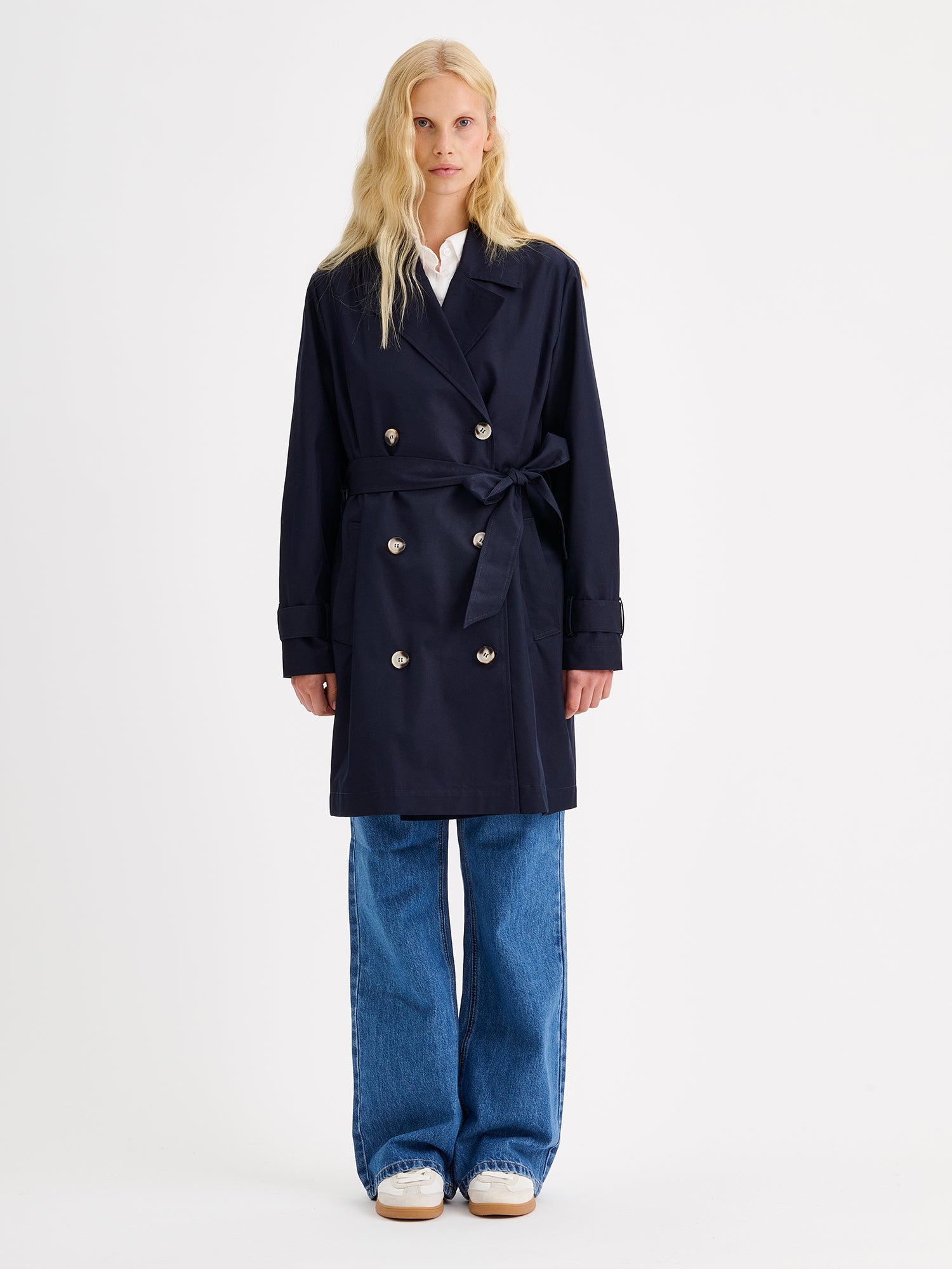 Nanson naisten tummansininen Midi Trenssi-takki vaaleahiuksisen mallin päällä farkkujen ja tennareiden kanssa.