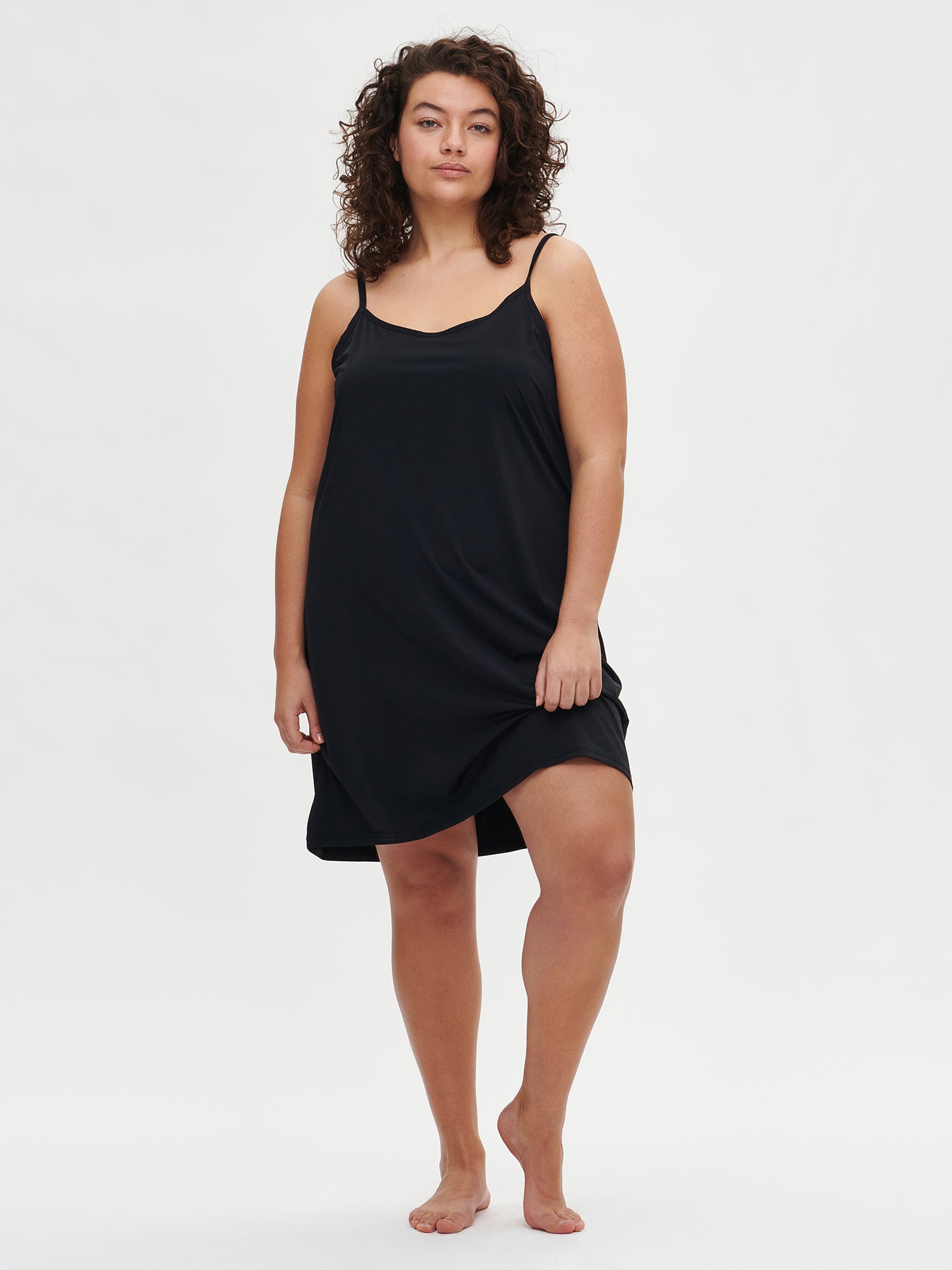 Nanson naisten musta Basic-alusmekko ohuilla olkaimilla, edestä kuvattuna plus size-mallin päällä. Helma kevyesti nostettu liikkeen näyttämiseksi.