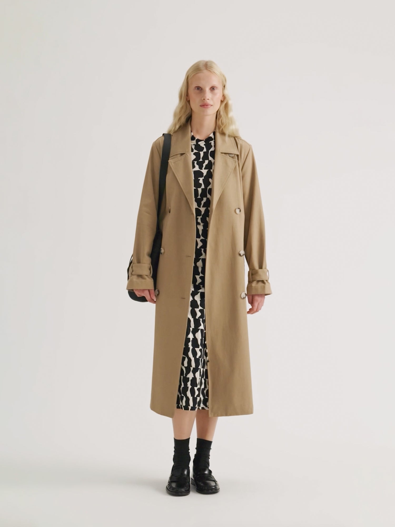 Naisten klassisen tyylikäs ruskea Klaara-trenssitakki mallin päällä. Trenssin alla Pieni Soraja-mekko ja olalla Naru Bucket musta nahkalaukku.