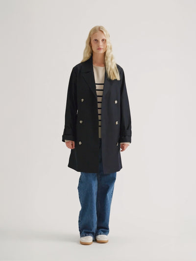 Nanson naisten tummansininen Midi Trenssi-takki vaaleahiuksisen mallin päällä farkkujen ja tennareiden kanssa, takki auki.